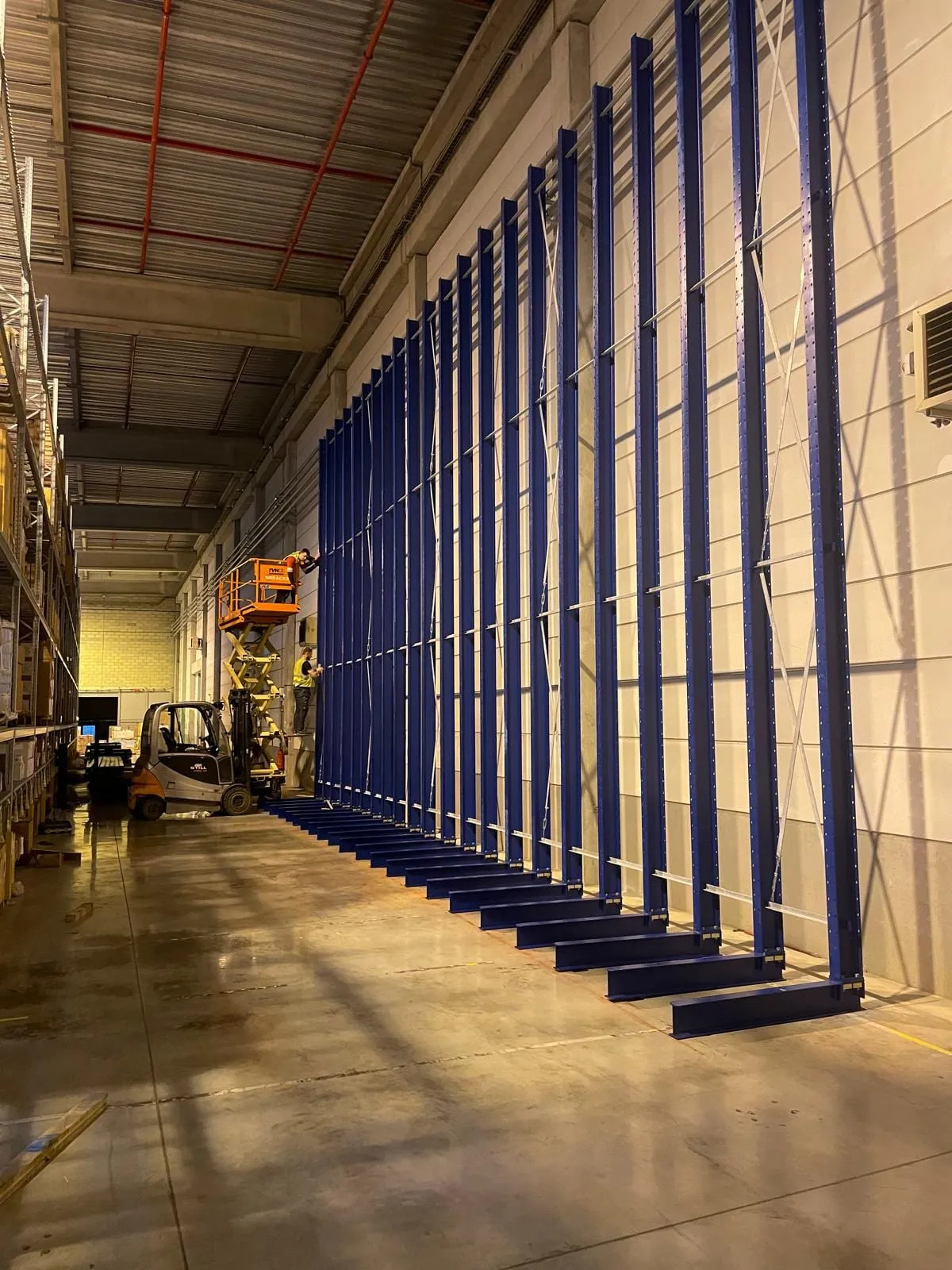 Rij blauwe staanders met schoren langs de magazijnwand, nog zonder armen, met achteraan een schaarlift aan het werk
