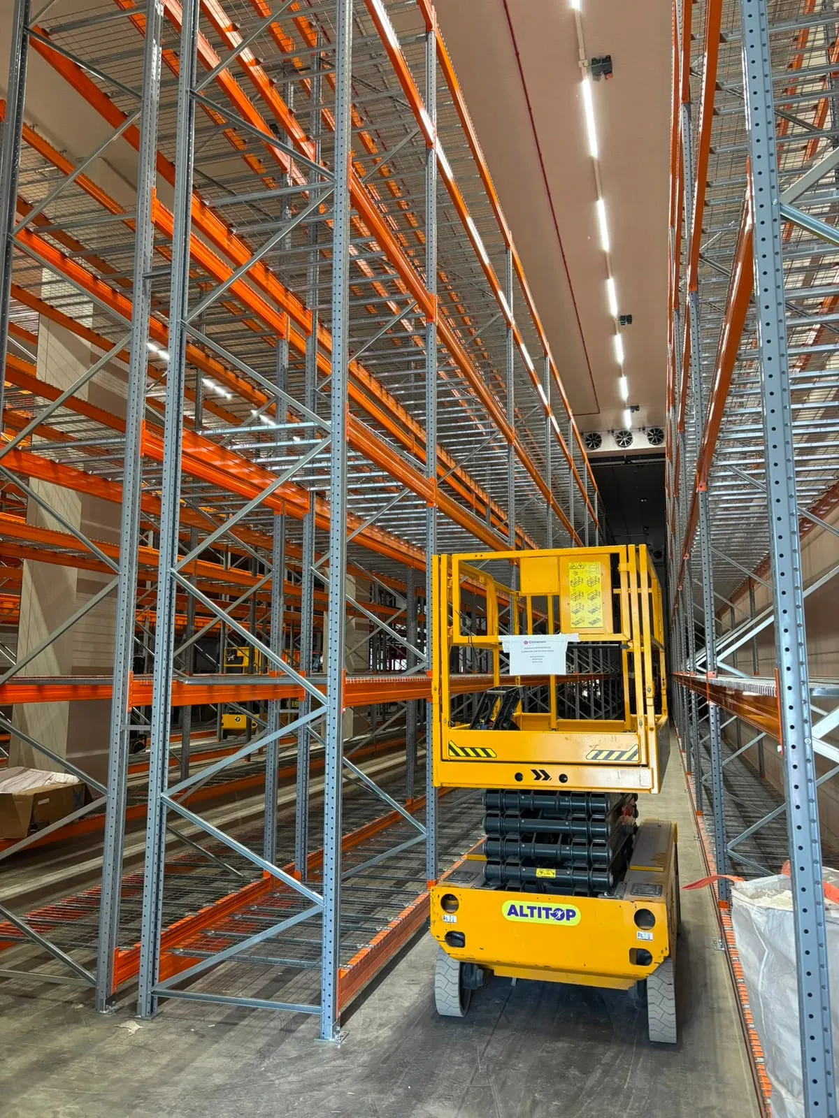 Hoge palletstellingen met grijze staanders, oranje liggers en gaaslegborden, met een schaarlift in de gang