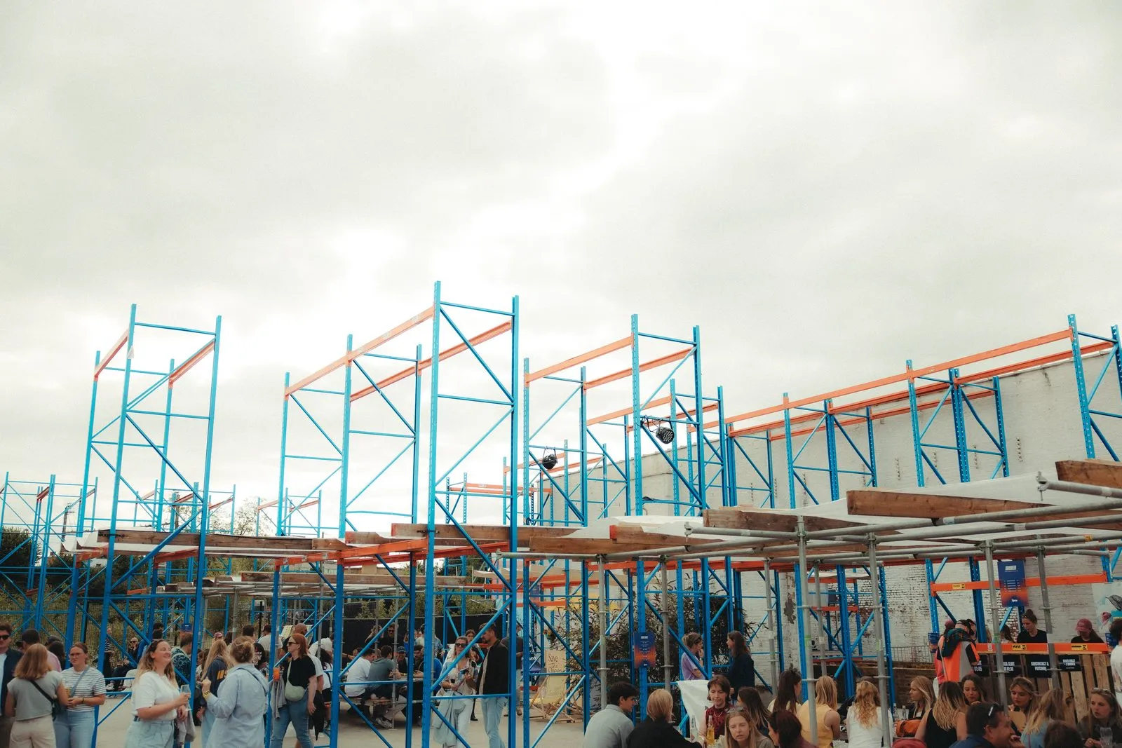 Blauwe staanders en oranje liggers rijzen als torens op boven festivalbezoekers, op een bewolkte namiddag