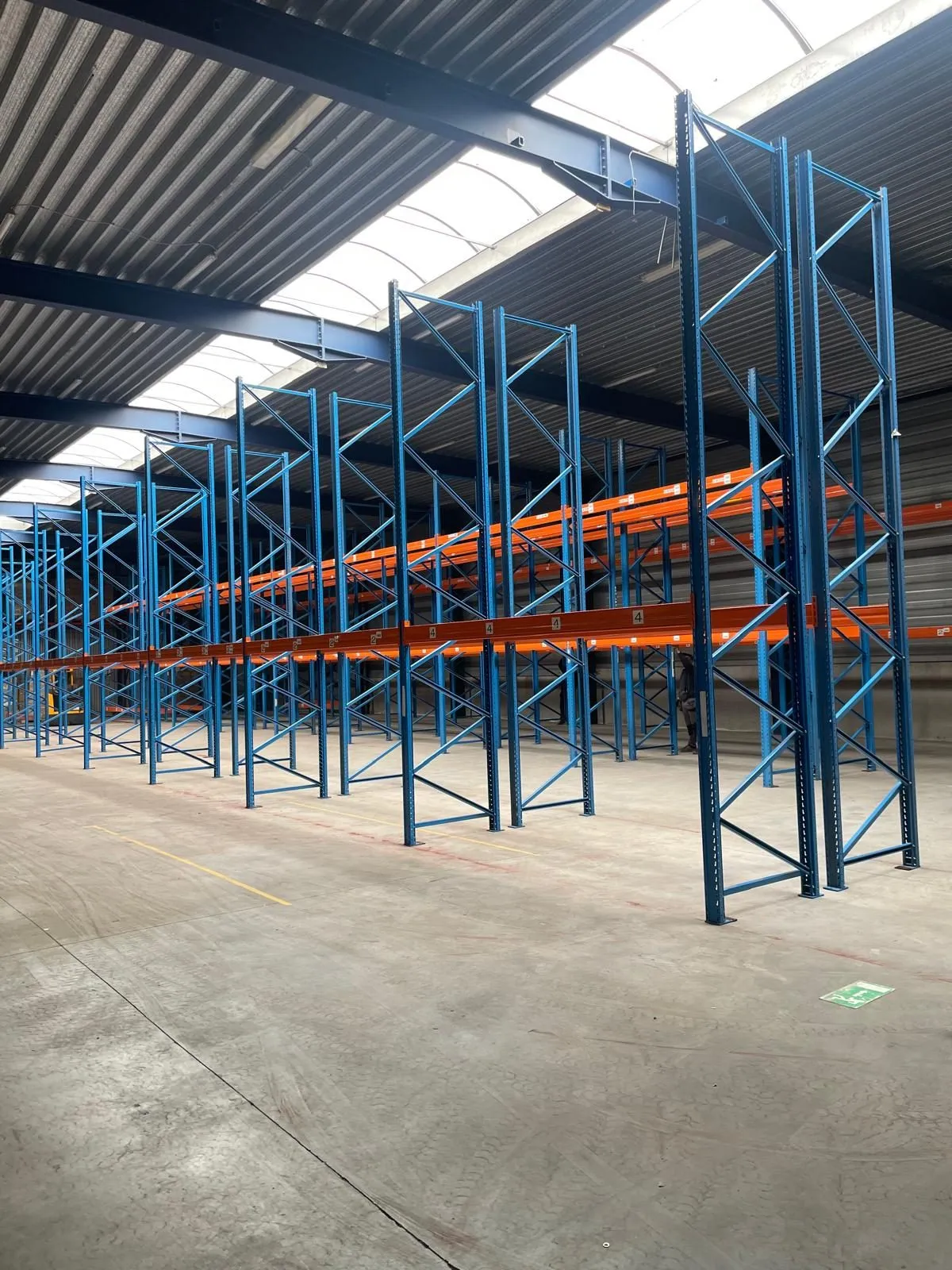 Rij palletrekken met blauwe staanders en oranje liggers in een hal met lichtstraten in het dak